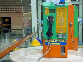 A brightly colored international payphone with orange, green, and yellow panels. It has a black handset attached with a metal cord and a numeric keypad with various instructions and buttons for operation. The phone is placed on a round marble surface with glass panels in the background displaying signs and reflections. The setting appears to be a public area, such as an airport or train station.