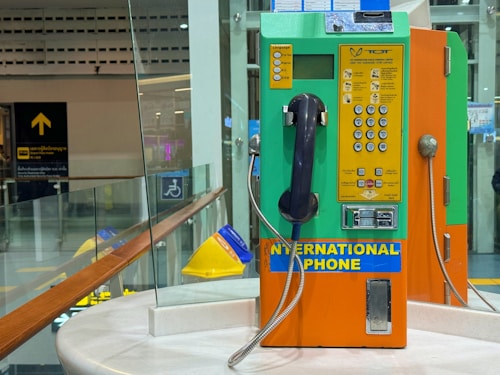 A brightly colored international payphone with orange, green, and yellow panels. It has a black handset attached with a metal cord and a numeric keypad with various instructions and buttons for operation. The phone is placed on a round marble surface with glass panels in the background displaying signs and reflections. The setting appears to be a public area, such as an airport or train station.