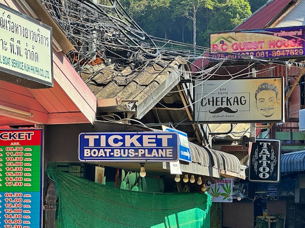 A bustling street scene with multiple storefront signs, including one for a ticket booth offering transportation services, a guest house, a tattoo parlor, and a shop associated with cheese. Several signs are in different languages, and electric cables hang loosely above. The area appears to be shaded with some greenery visible in the background.