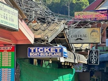 A bustling street scene with multiple storefront signs, including one for a ticket booth offering transportation services, a guest house, a tattoo parlor, and a shop associated with cheese. Several signs are in different languages, and electric cables hang loosely above. The area appears to be shaded with some greenery visible in the background.