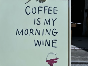 A sign with the phrase 'COFFEE IS MY MORNING WINE' written in bold black letters on a light-colored background. Below the text is a sketch of a wine glass with a dark red liquid, suggesting the humorous comparison between coffee and wine as morning rituals.