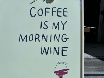 A sign with the phrase 'COFFEE IS MY MORNING WINE' written in bold black letters on a light-colored background. Below the text is a sketch of a wine glass with a dark red liquid, suggesting the humorous comparison between coffee and wine as morning rituals.