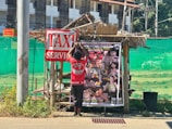 A person is hanging a poster on a small, rustic wooden structure covered by a thatched roof. The poster advertises an event, likely related to martial arts, with multiple fighters' images and the date December 23, 2023. A large sign nearby reads 'TAXI SERVICE'. The background includes a green mesh fence and a multi-story building with numerous windows.