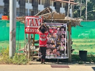 A person is hanging a poster on a small, rustic wooden structure covered by a thatched roof. The poster advertises an event, likely related to martial arts, with multiple fighters' images and the date December 23, 2023. A large sign nearby reads 'TAXI SERVICE'. The background includes a green mesh fence and a multi-story building with numerous windows.