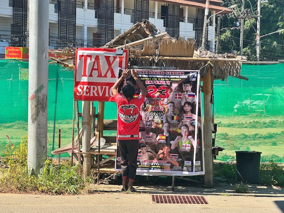 A person is hanging a poster on a small, rustic wooden structure covered by a thatched roof. The poster advertises an event, likely related to martial arts, with multiple fighters' images and the date December 23, 2023. A large sign nearby reads 'TAXI SERVICE'. The background includes a green mesh fence and a multi-story building with numerous windows.