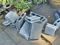 A stack of gray concrete blocks is placed on a paved surface surrounded by green potted plants and a large decorative pot. Some yellow leaves are scattered on the ground.