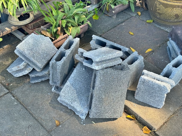 A stack of gray concrete blocks is placed on a paved surface surrounded by green potted plants and a large decorative pot. Some yellow leaves are scattered on the ground.
