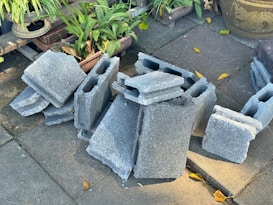 A stack of gray concrete blocks is placed on a paved surface surrounded by green potted plants and a large decorative pot. Some yellow leaves are scattered on the ground.