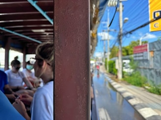 Several people are seated in a shaded area of a moving vehicle, possibly a bus or train, partially visible through a metal divider. The passengers are wearing casual clothing, with some using masks. The view outside shows a street scene with a sidewalk, electrical poles, and a billboard under a clear blue sky.