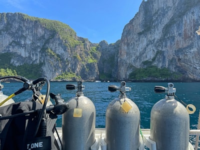 A scenic view featuring several scuba diving tanks on a boat with a backdrop of massive, rugged cliffs covered in patches of green foliage. The clear blue sky and calm water add to the tranquility of the scene.