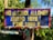 A weathered and colorful sign with blue and red background and yellow text that reads 'No alcohol allowed around here! Fine 1000 B.' It is mounted on a post surrounded by lush green foliage.