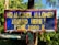 A weathered and colorful sign with blue and red background and yellow text that reads 'No alcohol allowed around here! Fine 1000 B.' It is mounted on a post surrounded by lush green foliage.