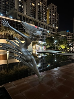 A modern sculpture of a stylized human figure with flowing hair is positioned at the edge of a reflective water surface. Tall buildings with many windows illuminate the background, creating a striking contrast with the night sky. The sculpture seems to be gracefully leaning forward, as if about to dive or dance.