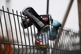 Several empty beverage cans are wedged into the wires of a metal fence, with some cans slightly crushed. The fence features a vertical and curving steel rod pattern, creating a modern industrial look.