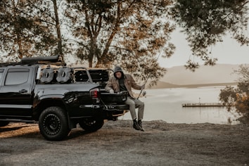 A person wearing a hoodie and jacket sits on the tailgate of a dark-colored pickup truck with camping gear. The scene is set by a lakeside with trees framing the view and mountains visible in the distance. The overall setting suggests a calm, outdoor recreational activity.