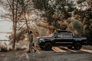 A person walks with a fishing rod near a black pickup truck parked on a dirt path surrounded by trees. Sunlight filters through the foliage creating a tranquil, early morning ambiance. The truck is equipped with off-road gear.