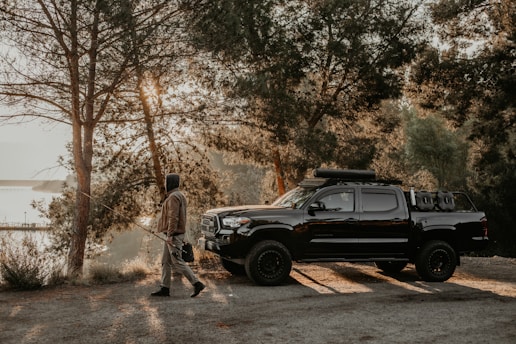 A person walks with a fishing rod near a black pickup truck parked on a dirt path surrounded by trees. Sunlight filters through the foliage creating a tranquil, early morning ambiance. The truck is equipped with off-road gear.