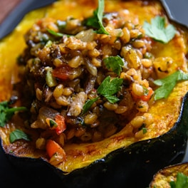 A roasted acorn squash filled with a colorful mixture of grains, vegetables, and herbs. The filling includes cooked grains, chopped vegetables like onions and tomatoes, and is garnished with fresh parsley. The squash is slightly charred around the edges and serves as an edible bowl for the vibrant stuffing.