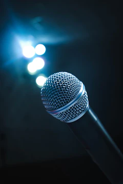 A close-up of a vintage microphone on stage with blue spotlight highlighting the drama masks in the background.