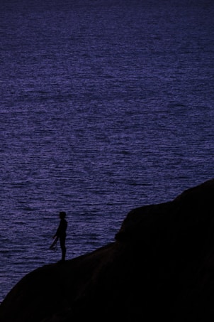 A silhouetted figure stands on a cliff edge overlooking a vast, dark blue ocean under low light conditions. The scene conveys a sense of solitude and contemplation.