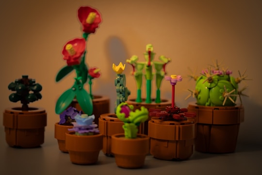 A collection of small, intricately built LEGO models depicting various types of plants and flowers, each placed in a brown pot. The arrangement includes a mix of floral shapes with vibrant colors, showcasing creativity and attention to detail. The scene is warmly lit, highlighting the craftsmanship of the miniature models.