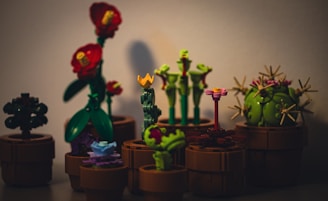 A collection of miniature plant figures crafted from colorful interlocking plastic bricks. The scene includes various types of plants, such as a cactus with spiky additions, flowers with vibrant blossoms, and other foliage. Each plant is placed in a brown pot and set against a plain background, creating a playful and imaginative display.