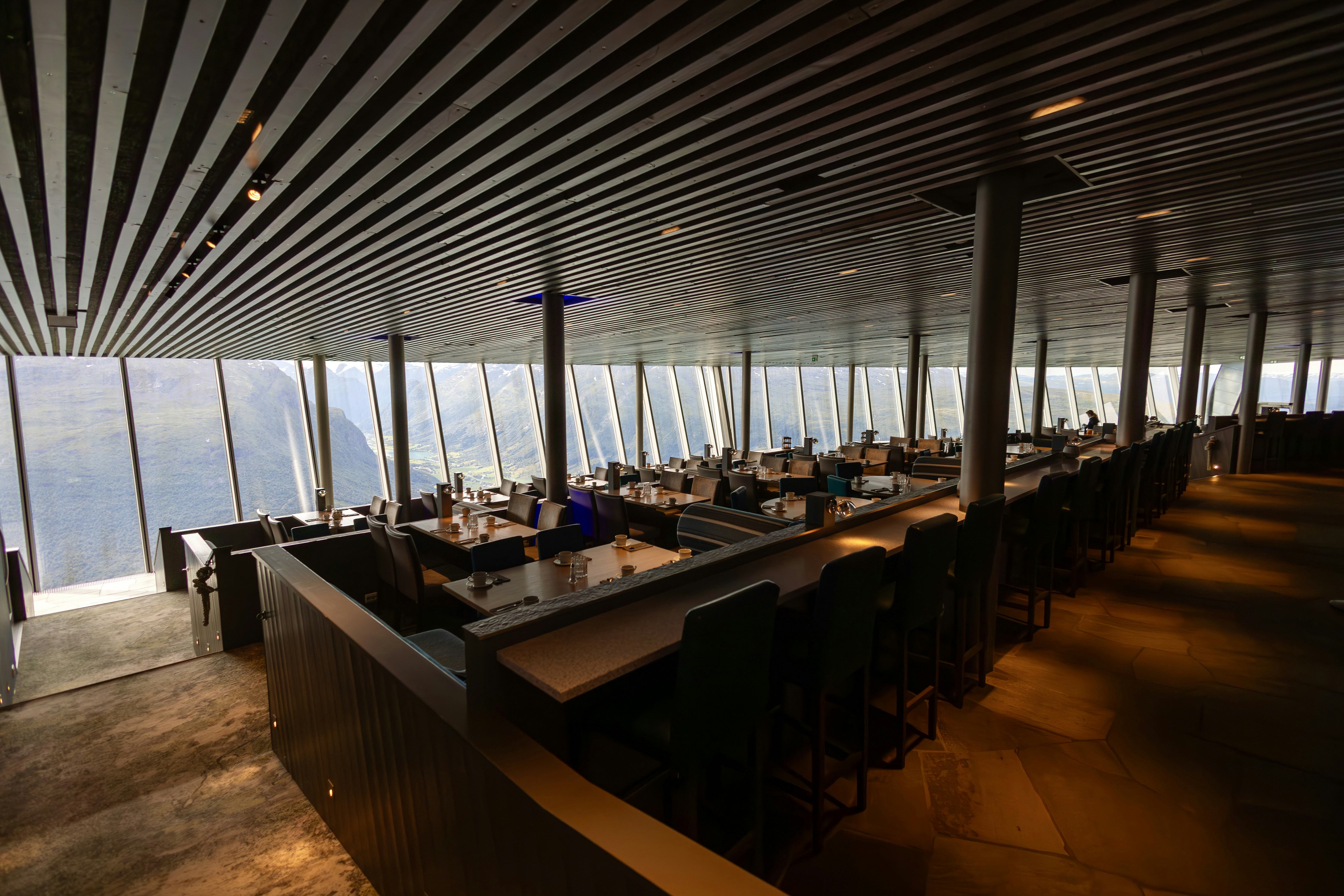 A modern restaurant interior with large floor-to-ceiling windows providing a scenic view of mountainous terrain. The ceiling features sleek, parallel lines that add to the contemporary design. The space is filled with neatly arranged tables and chairs, set for service.