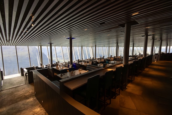 A modern restaurant interior with large floor-to-ceiling windows providing a scenic view of mountainous terrain. The ceiling features sleek, parallel lines that add to the contemporary design. The space is filled with neatly arranged tables and chairs, set for service.