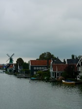 A scenic riverside village features traditional Dutch houses with steep roofs and a large windmill in the background. The houses have a distinct architectural style, with dark facades and white window frames. A small boat is moored by the riverbank, adding to the quaint rural setting. Lush greenery surrounds the area, and the sky is overcast, giving a sense of calm and tranquility.