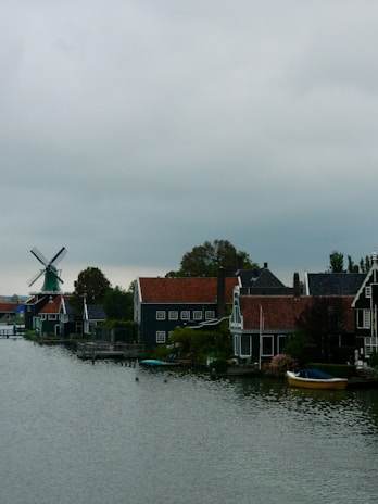 A scenic riverside village features traditional Dutch houses with steep roofs and a large windmill in the background. The houses have a distinct architectural style, with dark facades and white window frames. A small boat is moored by the riverbank, adding to the quaint rural setting. Lush greenery surrounds the area, and the sky is overcast, giving a sense of calm and tranquility.