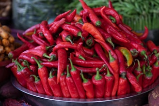A large pile of vibrant red chili peppers is arranged in a silver tray, with a few green peppers mixed in. Surrounding these are faint hints of additional vegetables, slightly out of focus, which suggests a market setting.