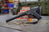 A black handgun with a suppressor is displayed on a surface surrounded by several boxes of Aguila ammunition. There are scattered bullets and an additional gun magazine lying nearby. The background appears to be an indoor setting possibly in a workshop or a store with shelves and various items slightly out of focus.