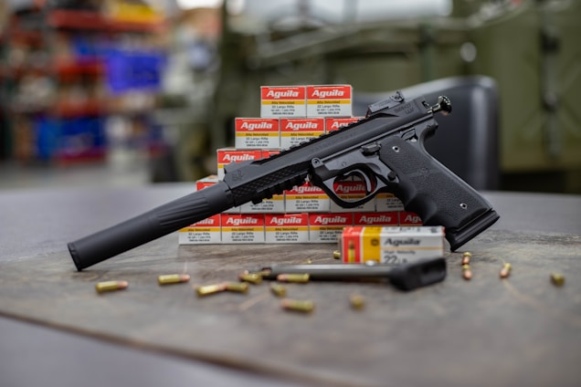 A black handgun with a suppressor is displayed on a surface surrounded by several boxes of Aguila ammunition. There are scattered bullets and an additional gun magazine lying nearby. The background appears to be an indoor setting possibly in a workshop or a store with shelves and various items slightly out of focus.