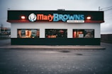 A fast-food restaurant with a prominent sign displaying 'Mary Browns' in bold letters. The building has large windows, some with promotional posters, and a lit 'open' sign. The exterior features a white facade with a black and red trim.