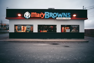 A fast-food restaurant with a prominent sign displaying 'Mary Browns' in bold letters. The building has large windows, some with promotional posters, and a lit 'open' sign. The exterior features a white facade with a black and red trim.