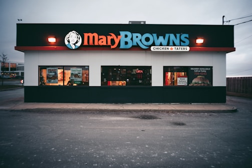 A fast-food restaurant with a prominent sign displaying 'Mary Browns' in bold letters. The building has large windows, some with promotional posters, and a lit 'open' sign. The exterior features a white facade with a black and red trim.