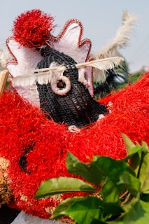 A person wearing a vibrant and elaborate traditional costume adorned with a bold red and black color scheme. The attire is decorated with feathers, netting, and textured materials. A prominent red pom-pom and white feathers are featured on the headpiece, while green leaves appear in the foreground.