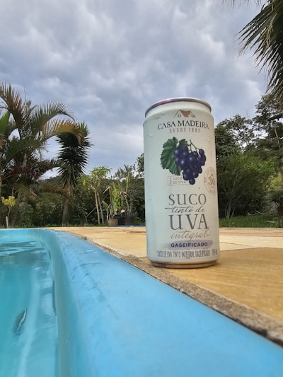 A can of Casa Madeira grape juice stands on the edge of a swimming pool. The pool water is a bright blue, contrasting with the can's design, which features an image of grapes. Behind the can, palm trees and other greenery are visible under a cloudy sky.