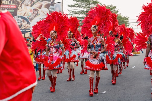 A group of people dressed in bright, colorful carnival costumes parade down a street. The outfits feature red feathers and sequins, and the participants wear coordinated shoes and headpieces. There are some trees in the background and a billboard displayed with various images.