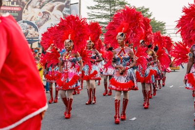 A group of people dressed in bright, colorful carnival costumes parade down a street. The outfits feature red feathers and sequins, and the participants wear coordinated shoes and headpieces. There are some trees in the background and a billboard displayed with various images.