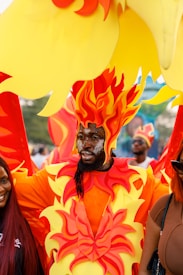 A person is wearing an elaborate costume that resembles flames, with vibrant red, yellow, and orange colors. The costume includes a headpiece that depicts fire, and the person's face has glitter makeup. They are flanked by others, one of whom has red hair, while the background shows more people and possibly additional costume elements.