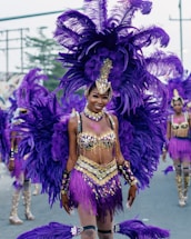 A vibrant parade participant dressed in an elaborate costume featuring large purple feathers, gold accents, and intricate beadwork. The person is smiling and surrounded by others wearing similarly colorful outfits, suggesting a carnival or festive setting.