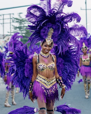 A vibrant parade participant dressed in an elaborate costume featuring large purple feathers, gold accents, and intricate beadwork. The person is smiling and surrounded by others wearing similarly colorful outfits, suggesting a carnival or festive setting.