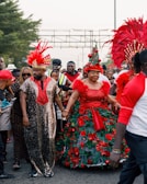 A festive parade with people wearing vibrant and elaborate costumes adorned with feathers and decorations. The scene is lively with a variety of individuals, some wearing sunglasses and hats, creating a cheerful atmosphere.