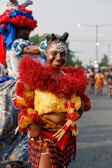 A person is dressed in vibrant traditional attire, featuring bright red, yellow, and black elements. The outfit includes intricate face paint and an elaborate headdress. In the background, other individuals are similarly dressed, participating in what appears to be a cultural celebration or festival.
