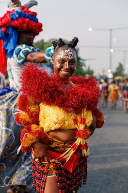 A person is dressed in vibrant traditional attire, featuring bright red, yellow, and black elements. The outfit includes intricate face paint and an elaborate headdress. In the background, other individuals are similarly dressed, participating in what appears to be a cultural celebration or festival.