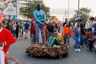 A street parade featuring performers in vibrant and colorful costumes, including a person wearing a blue outfit with a mask standing beside another person in a sitting position. The scene includes a large crowd of spectators, some taking photos, with trees and advertising billboards visible in the background.