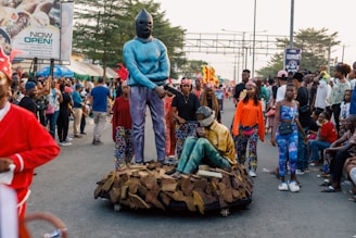 A street parade featuring performers in vibrant and colorful costumes, including a person wearing a blue outfit with a mask standing beside another person in a sitting position. The scene includes a large crowd of spectators, some taking photos, with trees and advertising billboards visible in the background.