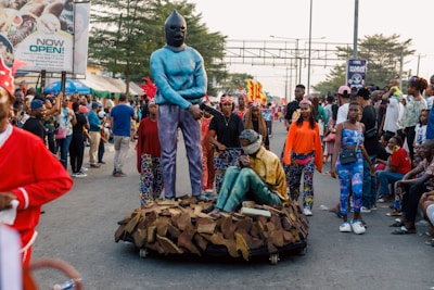 A street parade featuring performers in vibrant and colorful costumes, including a person wearing a blue outfit with a mask standing beside another person in a sitting position. The scene includes a large crowd of spectators, some taking photos, with trees and advertising billboards visible in the background.
