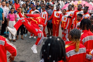 A person in vibrant red and checkered attire performs a dynamic leap in front of an audience. The crowd forms a supportive circle, dressed in matching costumes, suggesting a coordinated group performance. Spectators watch attentively, showcasing a lively and festive atmosphere.